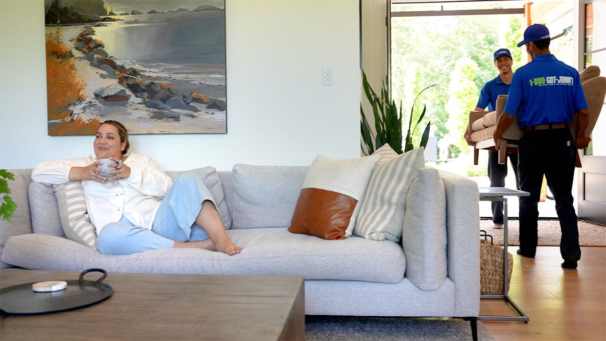 Relaxed woman on couch holding mug while two 1-800-GOT-JUNK? truck team members carry furniture out front door