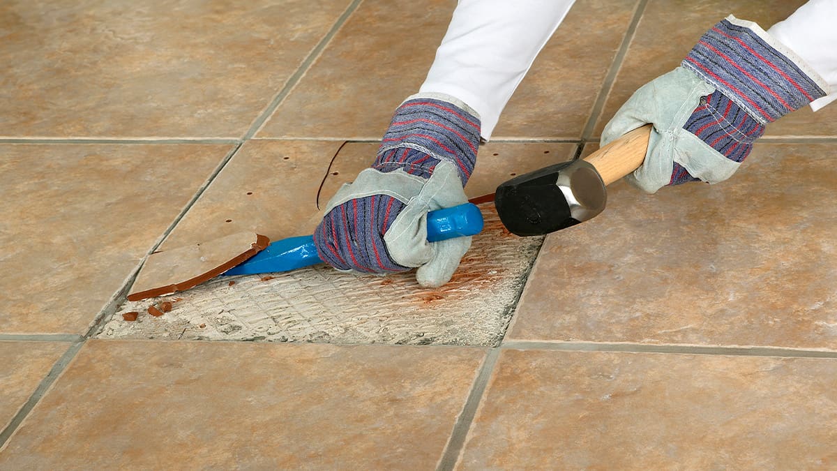 Gloved hands using a hammer and chisel to remove tile flooring