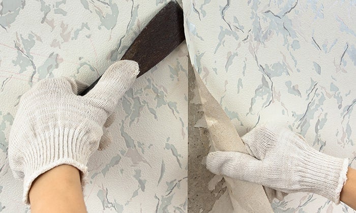 Gloved hands remove wallpaper using a scraper tool